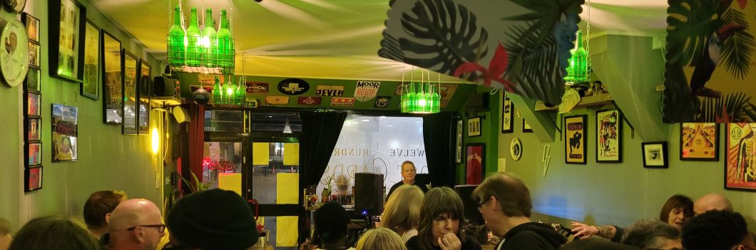 A view of the bar at 1200 Postcards, showing the tops of customers' heads and a DJ at the turntables in the background